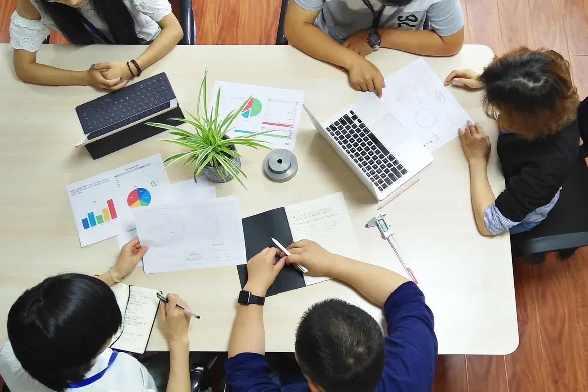 Overhead view of a team meeting, with laptops, technical drawings, charts and a caliper on the table